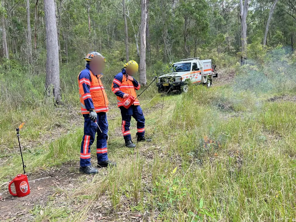 Two crew in turnout gear beside a 4x4 appliance, watching a small burn in open bush.