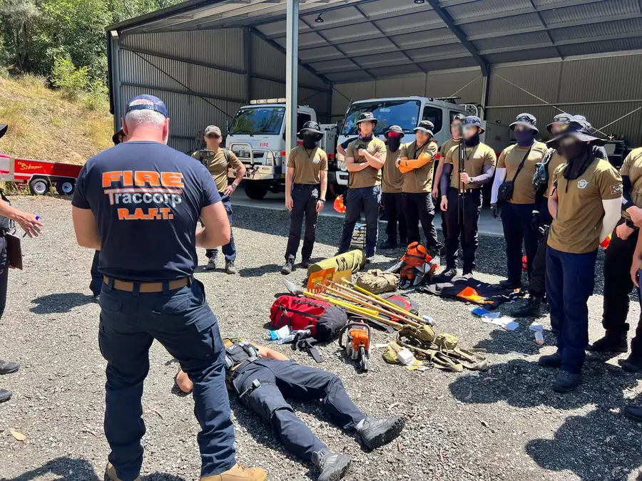 An instructor briefing international wildfire participants over equipment laid out on the ground, appliances in the bay behind them.